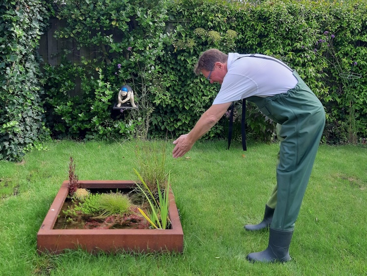 Een man in groene overall staat in zijn tuin en wijst naar planten in een border.
