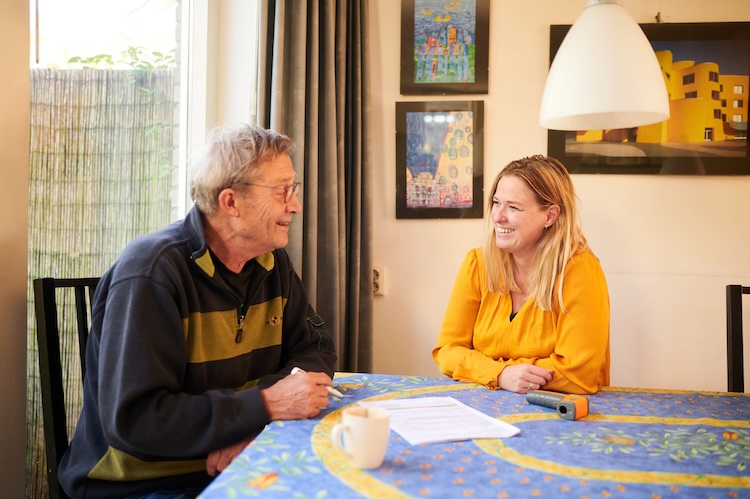 Twee mensen zitten in een woonkamer rond een tafel en kijken lachend naar elkaar.