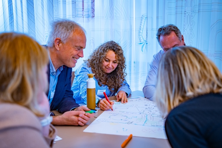 Een groep mensen zit rond een tafel en zij tekenen samen op een groot vel papier.