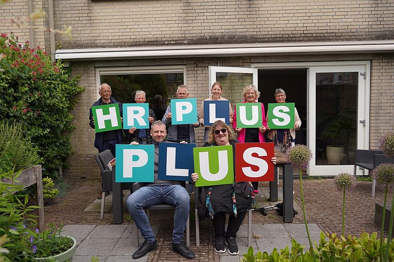 Zeven leden van het buurtteam Lisse houden, samen met buurtbegeleider Isabel van Buurkracht, kleurrijke letters vast. De letters lezen 'HR PLUS PLUS'.