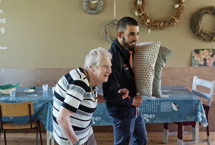 Een jong persoon heeft twee kussens onder zijn arm en helpt een oudere mevrouw met lopen in een huiselijke setting.