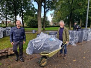 Twee mensen staan in een parkje, één persoon heeft een kruiwagen met daarin een regenton vast. Op de achtergrond staan nog meer regentonnen.
