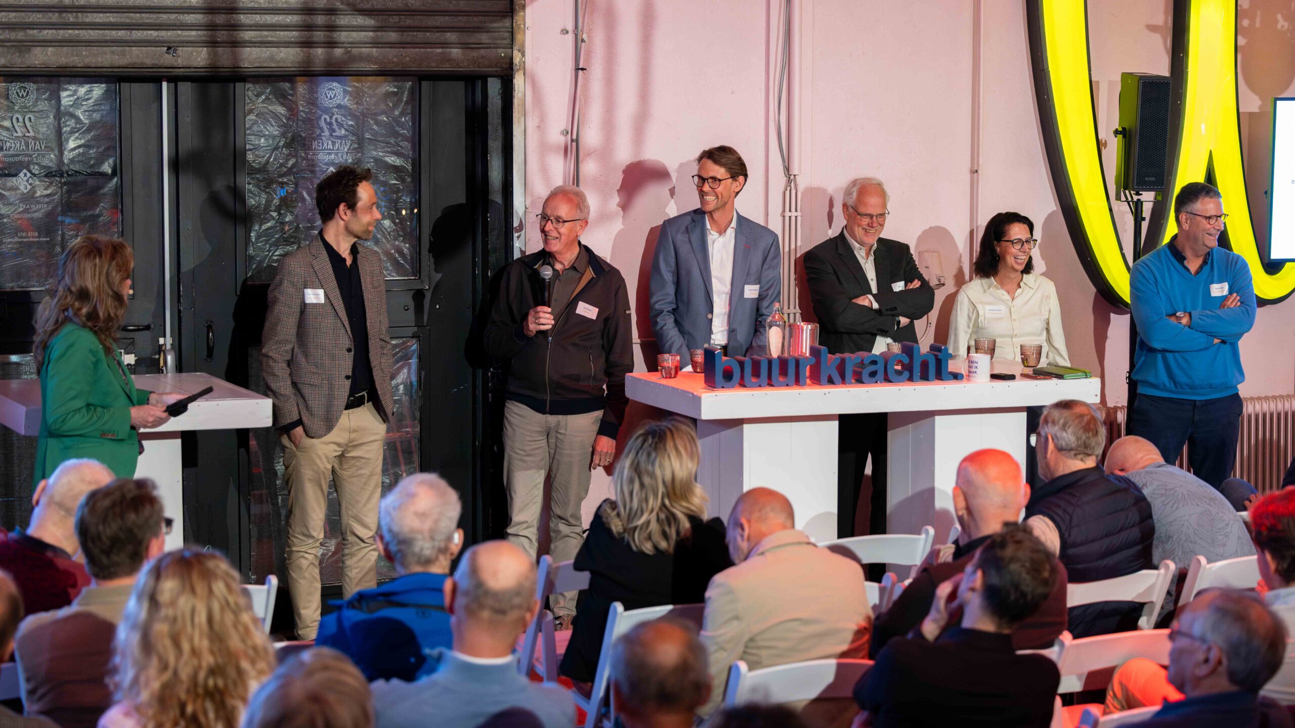 Een groep ambtenaren en bewoners staan voor een volle zaal met bezoekers tijdens het Samen sterk met buurkracht 2025 evenement. Ze zijn in gesprek en kijken lachend de zaal in.