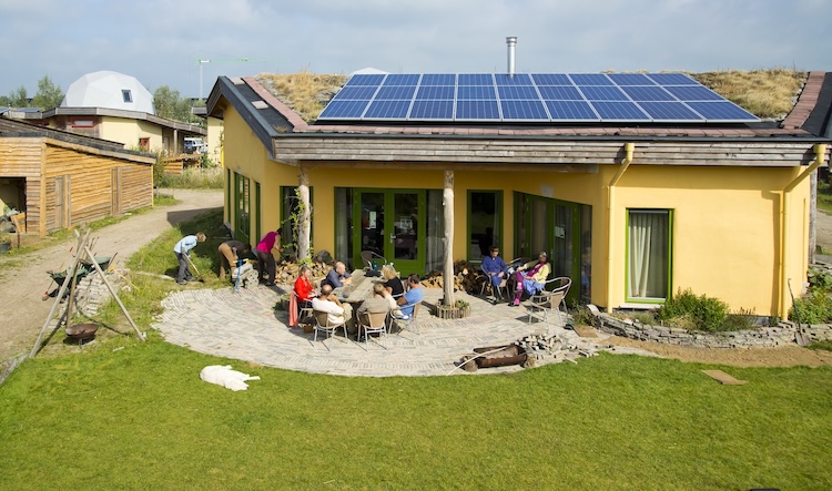 Een groep mensen zitten op een terras voor een groot huis met veel zonnepanelen op het dak.