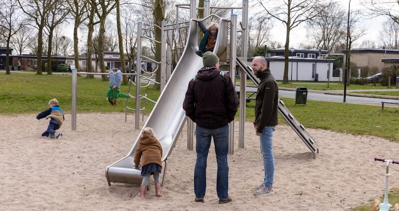 Twee mensen staan voor een speeltoestel te praten terwijl vier kinderen hierop aan het spelen zijn.