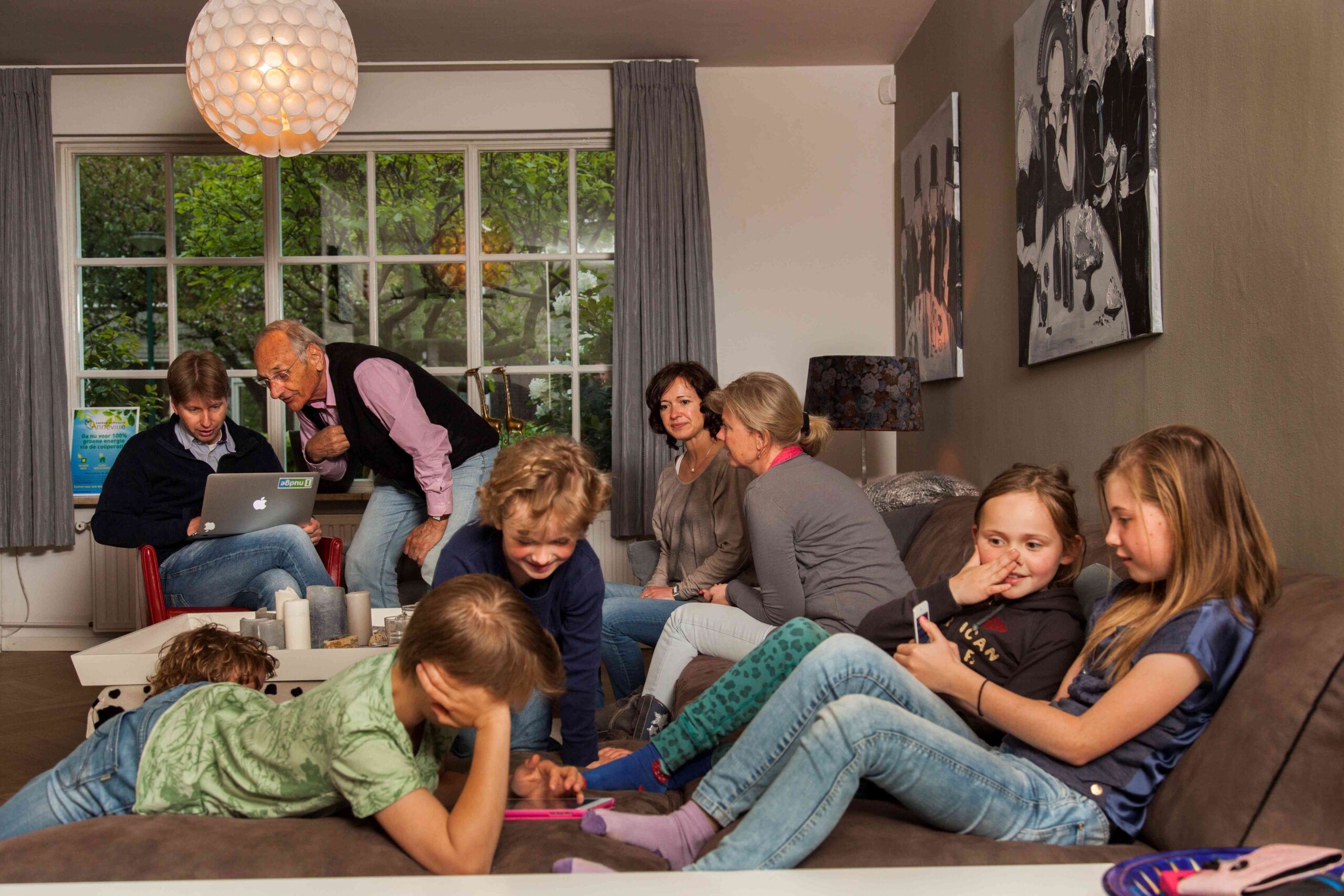Een groot gezin zit in de woonkamer van een huis. De ouderen praten met elkaar en de kinderen spelen op hun telefoons.
